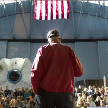 Man with red jacket and cap stands on stage with back towards camera, audience slightly below