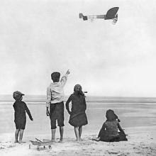 Black and white photo of four children on a beach. One points to an airplane in the distance.