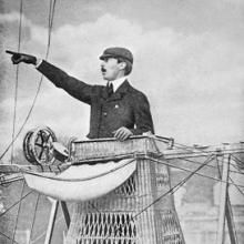 A man in basket-like airship gondola pointing to the left.