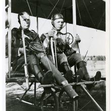 Two men in the seat of a Wright airplane. The man on the left is holding a large machine gun and the man on the right is at the controls.