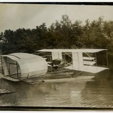 A biplane with an oval structure on the back floating on water with trees in the background.