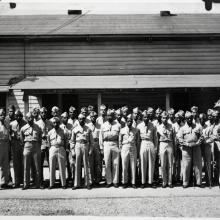 Black and white photo of approximately 30 Black men in uniform, posing in front of a wood building with an awning.