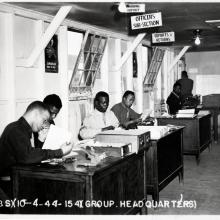 A line of four Black men work seated at a line of desks with boxes of paper and office equipment in front of them. Overhead is a sign that reads: "Officers Subsection."