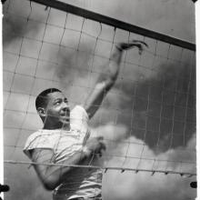 Black and white photo of a black man in a white tee shirt behind a volleyball net with his left hand in the process of spiking a ball (ball not visible)