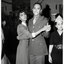 Black and white photo of two Black people dancing - (left) woman with wavy hair to just below the shoulders and bangs is turned sideways (right)  man with cropped hair holds her right hand with his left (and right arm around the her back).  Woman on right in dark dress with decorative belt is cut off