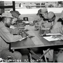 A group of Black men sit on the left and right side of a vertically place table.  They are all in uniform with various caps.  Each holds a notebook, pen, and is seated in front of a piece of a telegraph system.