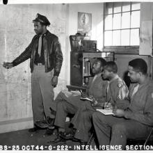 Black and white photograph. The entire back wall is a map of Europe.  A Black man in a jacket, hat, and tie points to the map with his right hand.  Three men sit in chairs to his right with notebooks and pens in hand.