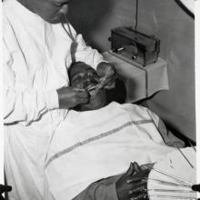 Black and White photo of a Black man standing with his dental tools in the open mouth another man, whose hands are clasped over his chest.  A tray of dental tools sits in the lower right foreground. 