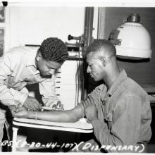 Black and white photo.  Black man on the left in a light uniform is holding a hypodermic needle to the right arm of a seated man, who is supporting his arm with his right hand.