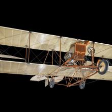 Khaki and orange colored biplane craft suspended in air against a black background.