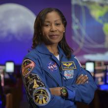 Stephanie Wilson stands with her arms folded, wearing a blue jacket with many space mission patches.