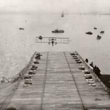 A Curtiss Model D flying toward a ship which has a makeshift runway. Other boats dot the water.