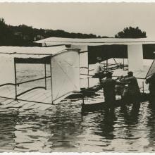 One-half right rear view of the a Blériot floatplane on the water. Men stand in the water next to it.