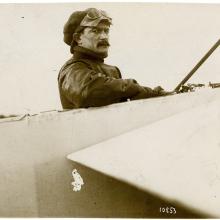 A man wearing a hat and goggles on his head, sitting in the cockpit of an airplane, looking toward the camera.