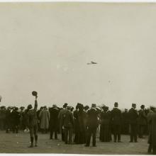 A crowd of people, some cheering, looking toward the sky where an airplane can be seen in the distance.