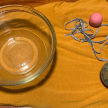 A photograph of supplies for an activity including a bowl, a ball that looks like Earth, another ball, a blanket, and a string. 