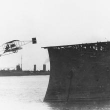 An old photograph of a biplane taking off a boat. 