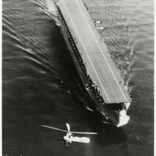 Black and white grainy image of USS Langley in water and an aircraft flying around it