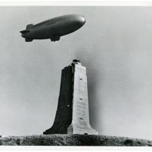 One-half right front view of a U.S. Navy airship in flight over the square column-shaped Wright brothers memorial, which has both brothers names inscribed on it.