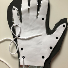 A cut out of a hand on printer paper with hole punches around the outside, stacked on top of a cut out of a hand on Mylar, stacked on top of a cut out of a glove on foam. A piece of yarn with tape around the end creating a point is threaded through the first hole.