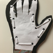 A cut out of a hand on printer paper with hole punches around the outside and yarn threaded through the holes, stacked on top of a cut out of a hand on Mylar, stacked on top of a cut out of a glove on foam. 