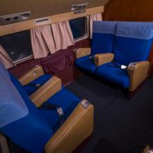 A quartet of four seats in sets of two facing each other. Behind the seats is a row of windows with curtains. The seats are blue and the surrounding walls brown. 