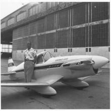 A standing person poses next to a small monoplane in front of an airplane hangar. 