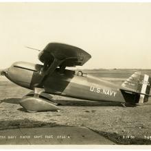 Sepia-toned photo of the left side view of a person in a Curtiss racer monoplane on the ground. The aircraft has U.S. Navy written on the side. 