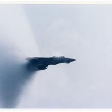 A jet aircraft emerging from a cone shaped cloud of vapor in the air. 