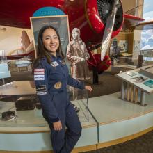 A woman in a flight uniform poses next to a bright colored early aircraft in a museum setting.