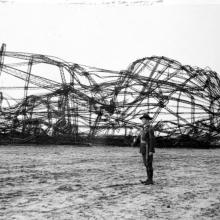 A person in uniform stands next to the wreckage of an airship.