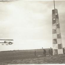 Farman-type biplane rounds a pylon in a field as a handful of people look up, some waving their hats in the air.