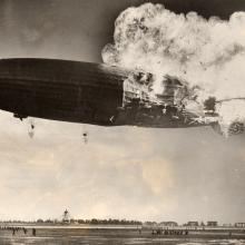 An airship is seen with the back half of its body on fire as it is still in the air.