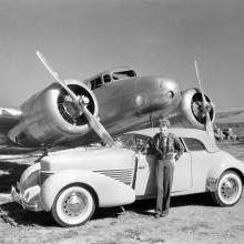 A wideshot of a woman standing next to a car with a aircraft in the background.