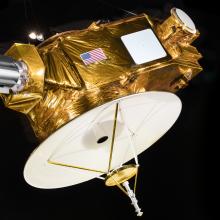 A spacecraft with a round disk on the bottom, a cylinder on the side, and a golden body with the United States flag on it.  