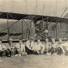 Nine people sit in a row on the ground, another person stands behind behind them in the center. They pose in front of a biplane.