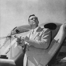 Black and white image of a man standing in front of an airplane, holding an object in his hand, and looking to the right.