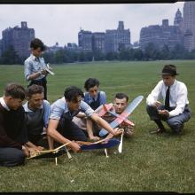 A group of six young men of various ages gather around three model airplanes in a park with a city skyline in the distance. To the right is an adult crouching on the ground next to them with what seems to be either a stopwatch or notebook. 