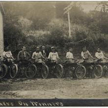 Eleven people pose with motorcycles in front of a building and bushes in this sepia-toned photo.