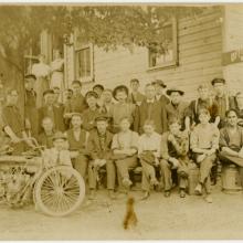 A large group of people pose in front of a building in this sepia-toned photo. A motorcycle is propped in front of the left side of the group.