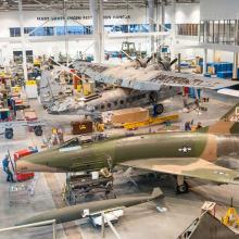 An airport hanger with two planes on the ground. They are surrounded by equipment to work on them.