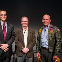 Three men smile at a camera, one wears a flight jacket. 