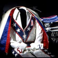 A red, white, and blue colored jumpsuit and cape propped in a sitting position. In the background is a partial view of a red, white, and blue motorcycle.