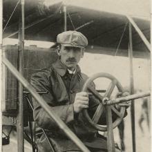 A man sits behind the steering wheel of a biplane, looking toward the camera. The photo looks old and is dated August 1909.