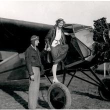 A woman looks toward the camera as she climbs through the door of an aircraft. A man to the left, Clyde Cessna, assists her.