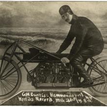 A man sits posed on a motorcycle, looking at the camera. He is in front of what is presumably a fake background meant to look like the beach.