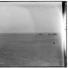 A photo in which a few small buildings can be seen in the distance. In the foreground is a flat plain. Just beyond the buildings the ocean can be seen.