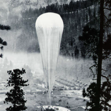 A black and white photograph of a balloon inflating above the trees. 