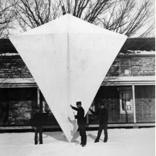 A giant four sided kite. Several men stand in front of the kite, and it is more than twice as tall as they are. 