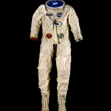 A cream colored space suit with accents of red and blue posed against a black background. There are boots attached but no helmet or gloves.